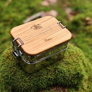 Pode incluir: Uma lancheira de madeira com um recipiente de metal. A tampa é gravada com o texto "Maggi" e "Petra".