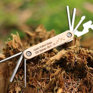 May include: A multi-tool with a wooden handle engraved with the text "Gute Fahrt Papa Wir lieben dich" and a mountain design. The tool has multiple attachments, including a screwdriver, a bottle opener, and a wrench.