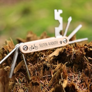 Op de afbeelding: Een multitool met een houten handvat en diverse metalen hulpstukken. Het handvat is gegraveerd met een fietsafbeelding en de tekst "SELTEN RADLOS!". De tool is gedeeltelijk open en toont verschillende functies.