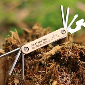 Może przedstawiać: Wielofunkcyjne narzędzie z drewnianym uchwytem grawerowanym tekstem "Gute Fahrt Papa Wir lieben dich" i wzorem gór. Narzędzie ma wiele dodatkowych elementów, w tym śrubokręt, otwieracz do butelek i klucz.