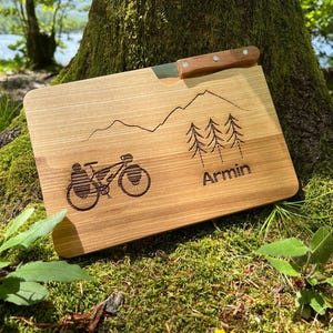Può includere: Un tagliere in legno con una fessura per il coltello, inciso con una scena di montagna, una bicicletta e il nome "Armin".