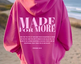 May include: A vibrant pink hoodie with the words "MADE FOR MORE" printed on the back in a lighter pink. Below the text is a Bible verse. The hoodie is worn by a person standing on a beach.