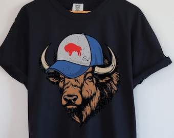 May include: Black t-shirt with a graphic of a bison wearing a blue and white baseball cap with a red bison logo. The bison has brown fur and large horns. The shirt is displayed on a wooden hanger.