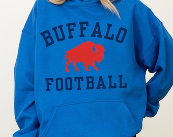 Könnte beinhalten: Ein blauer Kapuzenpullover mit den Worten "BUFFALO FOOTBALL" in marineblauen Blockbuchstaben. Eine rote Büffelgrafik ist zwischen den Wörtern zentriert. Der Pullover hat eine Vordertasche und lange Ärmel.