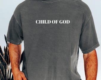 May include: A gray t-shirt with the text "CHILD OF GOD" in white block letters. The shirt is a relaxed fit and has short sleeves. The model is wearing blue jeans and a brown watch.