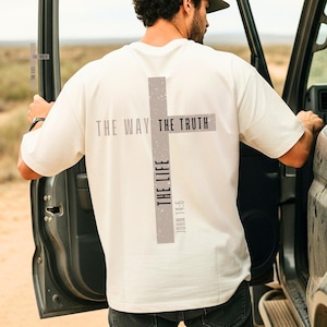 May include: An ivory short-sleeved t-shirt featuring a large grey cross graphic on the back. The text "THE WAY," "THE TRUTH," and "THE LIFE" is printed across the cross, with "JOHN 14:6" positioned vertically below. The word "IVORY" appears at the top.