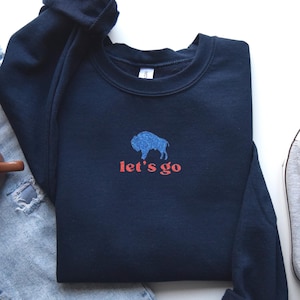 May include: A navy blue sweatshirt with a blue bison graphic and the words "let's go" in red. The sweatshirt is folded, with a pair of jeans, a mug, and a sneaker visible in the background.