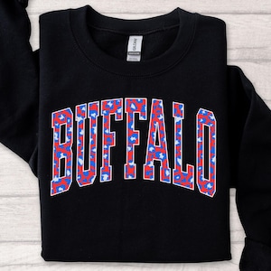 A black sweatshirt featuring bold arched “BUFFALO” varsity lettering filled with a bright red-and-blue patterned print and outlined in white. The vibrant design stands out sharply against the dark fabric, giving it a trendy, sporty Buffalo-pride look.