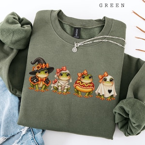 May include: A military green sweatshirt featuring a graphic of four frogs in Halloween costumes: a witch, a mummy, a hot dog, and a ghost. The text "MILITARY GREEN" is printed at the top. The sweatshirt has a crew neckline and long sleeves.