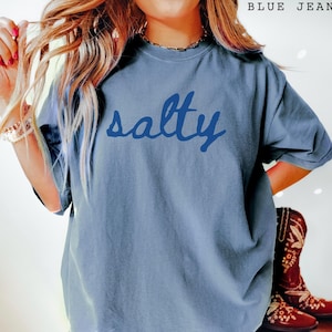 Blue Jean unisex graphic t-shirt featuring the word “salty” in bold navy handwritten script across the chest. Relaxed fit tee with a coastal, beach-inspired vibe, styled casually with layered jewelry and western boots for a trendy everyday look. Soft washed blue color makes it perfect for summer outfits, vacation wear, ocean lovers, and fun statement style.