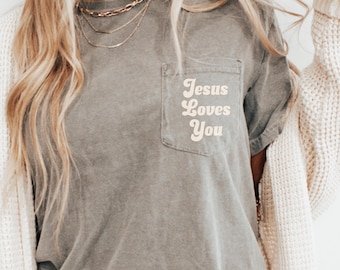 May include: A gray t-shirt with a pocket on the left side. The pocket has the text "Jesus Loves You" in a retro font.