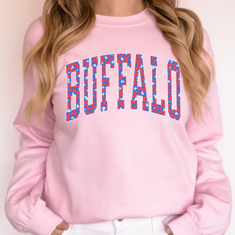 May include: A light pink sweatshirt with long sleeves. The word "BUFFALO" is printed across the chest in a bold, collegiate-style font. The letters are filled with a pattern of red and blue shapes.