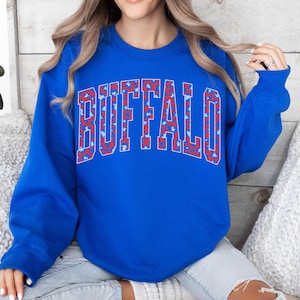 May include: Royal blue sweatshirt with the word "BUFFALO" in large, bold letters across the chest. The letters are outlined in red and filled with a red and white pattern. The sweatshirt has long sleeves and a crew neck.