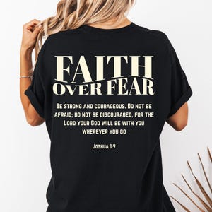 May include: Black t-shirt with the text "FAITH OVER FEAR" in a bold, off-white font. Below the main text is the phrase "BE STRONG AND COURAGEOUS..." and the Bible verse Joshua 1:9. The shirt is a casual style.