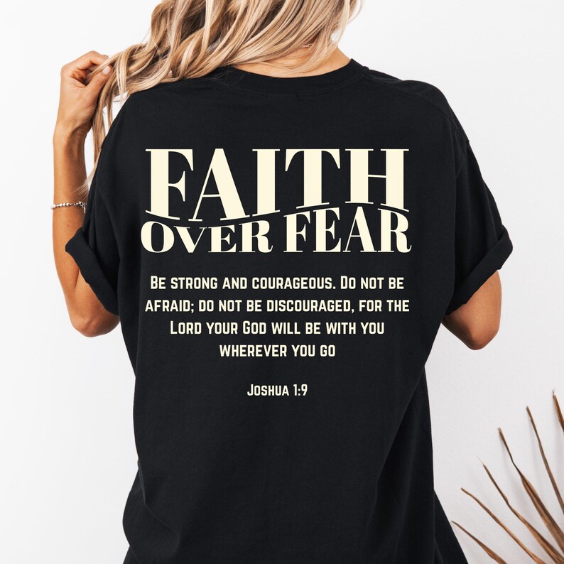 May include: Black t-shirt with the text "FAITH OVER FEAR" in a bold, off-white font. Below the main text is the phrase "BE STRONG AND COURAGEOUS..." and the Bible verse Joshua 1:9. The shirt is a casual style.