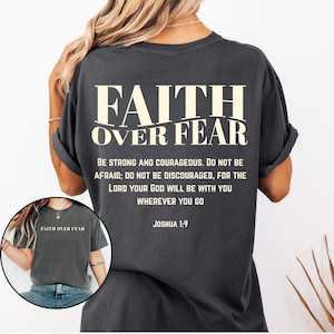May include: Dark gray t-shirt with the text "FAITH OVER FEAR" in a bold, off-white font. Below the main text is a quote from Joshua 1:9. The shirt has rolled-up sleeves and a relaxed fit.
