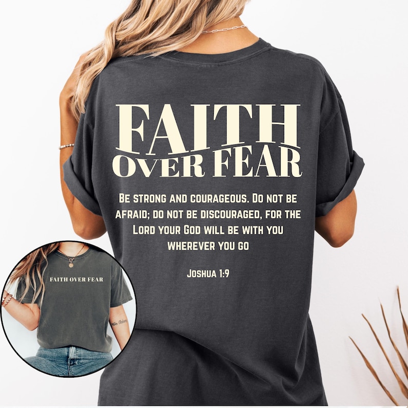 May include: Dark gray t-shirt with the text "FAITH OVER FEAR" in a bold, off-white font. Below the main text is a quote from Joshua 1:9. The shirt has rolled-up sleeves and a relaxed fit.