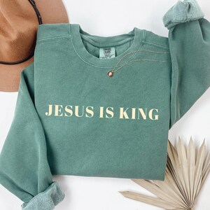 May include: A sage green sweatshirt with the text "JESUS IS KING" in a light beige color. The long-sleeved top has rolled cuffs and a crew neck. A gold necklace with a pendant rests on the shirt. A brown hat is visible in the background.