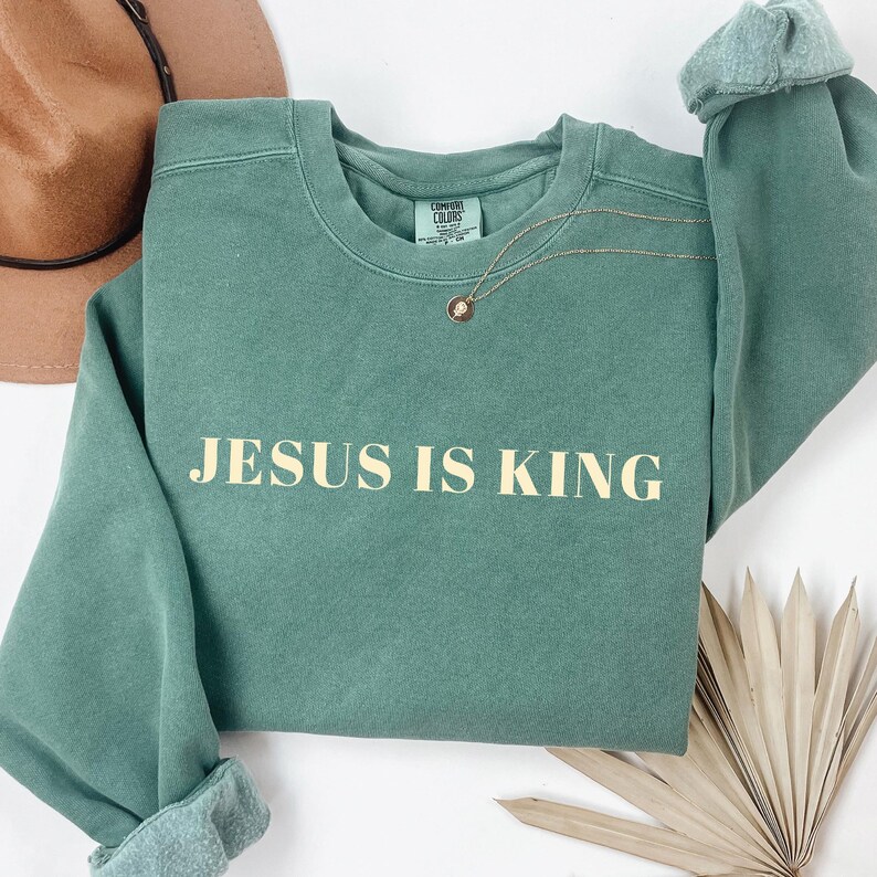 May include: A sage green sweatshirt with the text "JESUS IS KING" in a light beige color. The long-sleeved top has rolled cuffs and a crew neck. A gold necklace with a pendant rests on the shirt. A brown hat is visible in the background.