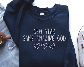 May include: A navy blue sweatshirt featuring the white text "NEW YEAR SAME AMAZING GOD" and three heart outlines. The garment is folded, with parts of jeans and a trainer visible in the background.