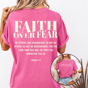May include: A pink t-shirt with the words "FAITH OVER FEAR" printed in white on the back. Below the text is a Bible verse from Joshua 1:9. The shirt is a short-sleeved, casual style.