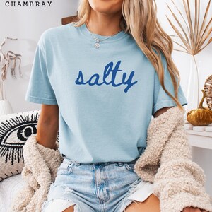 以下が含まれることがあります： 濃い青色のスクリプトで「salty」という言葉が書かれた水色のTシャツ。Tシャツは半袖でクルーネックです。画像左上には「CHAMBRAY」の文字がプリントされています。