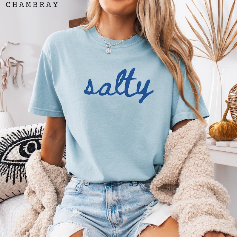 以下が含まれることがあります： 濃い青色のスクリプトで「salty」という言葉が書かれた水色のTシャツ。Tシャツは半袖でクルーネックです。画像左上には「CHAMBRAY」の文字がプリントされています。