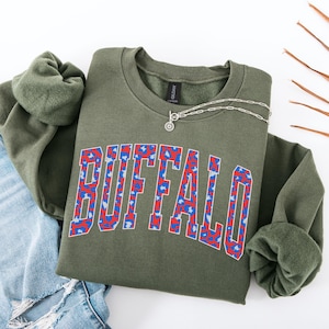 May include: An olive green sweatshirt with the word "BUFFALO" in a bold, collegiate-style font. The letters are filled with a red and blue leopard print pattern. A silver necklace with a sunburst pendant rests on the shirt.