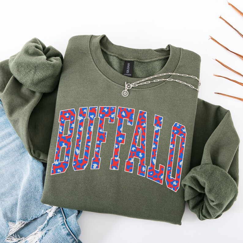 May include: An olive green sweatshirt with the word "BUFFALO" in a bold, collegiate-style font. The letters are filled with a red and blue leopard print pattern. A silver necklace with a sunburst pendant rests on the shirt.