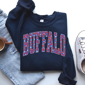 A navy sweatshirt featuring bold arched “BUFFALO” varsity letters filled with a red-and-blue abstract pattern and outlined in white. The design pops against the dark fabric, giving it a sporty, trendy Buffalo-pride look.