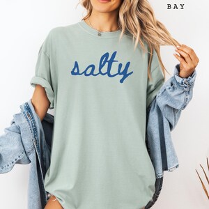 以下が含まれることがあります： 水色のTシャツに青い筆記体で「salty」の文字。Tシャツは半袖でゆったりとしたフィット感。モデルはTシャツの上にデニムジャケットを着ています。