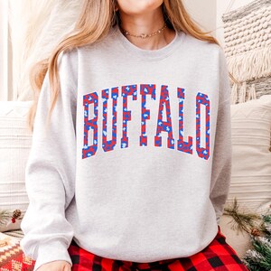 May include: A light gray sweatshirt with the word "BUFFALO" in large, bold letters across the chest. The letters are red with a blue and white star pattern. The sweatshirt is a casual style.