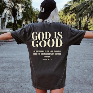 May include: A black t-shirt with the text "GOD IS GOOD" in white lettering. The shirt also features a quote from Psalm 107:1 in smaller white lettering.