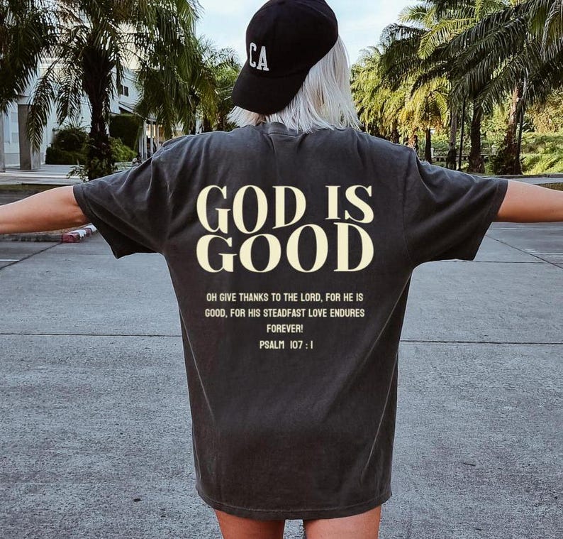 May include: A black t-shirt with the text "GOD IS GOOD" in white lettering. The shirt also features a quote from Psalm 107:1 in smaller white lettering.