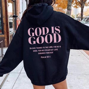 May include: Black hooded sweatshirt with the text "GOD IS GOOD" in pink lettering on the back. The text "OH GIVE THANKS TO THE LORD, FOR HE IS GOOD, FOR HIS STEADFAST LOVE ENDURES FOREVER! PSALM 107:1" is printed below in smaller lettering.