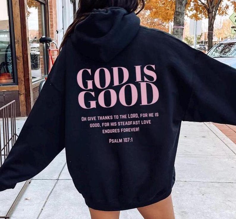May include: Black hooded sweatshirt with the text "GOD IS GOOD" in pink lettering on the back. The text "OH GIVE THANKS TO THE LORD, FOR HE IS GOOD, FOR HIS STEADFAST LOVE ENDURES FOREVER! PSALM 107:1" is printed below in smaller lettering.