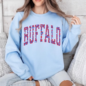 A light blue sweatshirt featuring bold arched “BUFFALO” varsity lettering filled with a red-and-blue abstract pattern and outlined in white. Trendy, sporty, and bright—perfect for showing Buffalo pride in a fun, modern style.