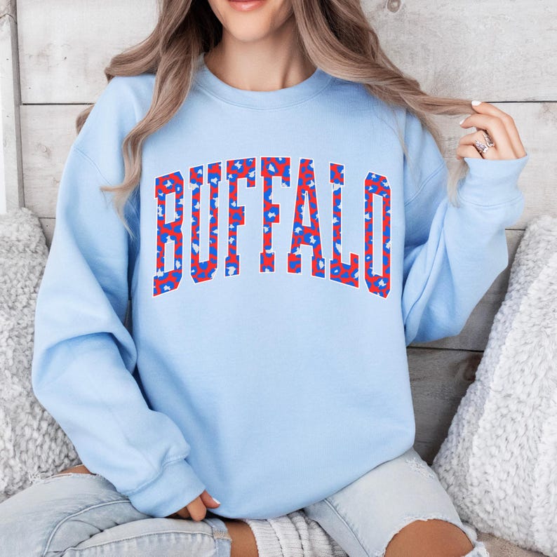 A light blue sweatshirt featuring bold arched “BUFFALO” varsity lettering filled with a red-and-blue abstract pattern and outlined in white. Trendy, sporty, and bright—perfect for showing Buffalo pride in a fun, modern style.