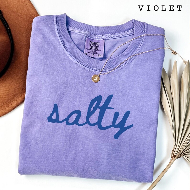 Violet Comfort Colors unisex graphic t-shirt featuring the word “salty” in bold blue handwritten script across the front. Soft washed purple tee folded flat with simple jewelry and neutral styling accents for a clean, coastal-inspired look. Relaxed fit and minimalist statement design, perfect for summer outfits, beach days, vacation wear, or ocean lovers who enjoy fun graphic tees.