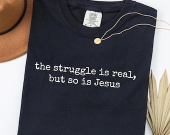 Puede incluir: Una camiseta azul marino con el texto blanco "the struggle is real, but so is Jesus". Un collar dorado con un pequeño colgante descansa sobre la camiseta. Un sombrero marrón es visible en la esquina superior izquierda.