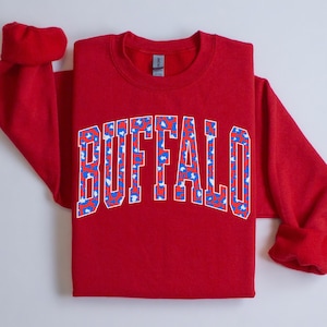 May include: A red sweatshirt with the word "BUFFALO" in large, white, block letters. The letters are filled with a pattern of blue and red spots. The sweatshirt has long sleeves and a crew neck.