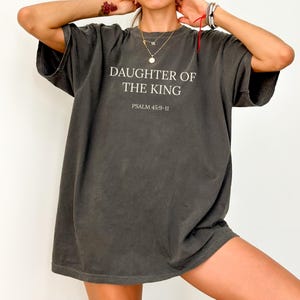 May include: Dark grey oversized t-shirt with the text "DAUGHTER OF THE KING" and "PSALM 45:9-11" printed in white. The shirt has short sleeves and a relaxed fit, suitable for casual wear.