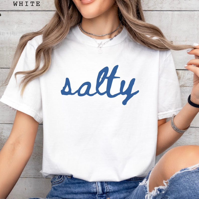 以下が含まれることがあります： 青い筆記体で「salty」と書かれた白いTシャツ。Tシャツは半袖でクルーネックです。画像左上には「WHITE」の文字がプリントされています。