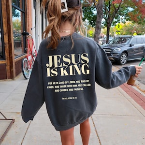 May include: Dark gray sweatshirt with the text "JESUS IS KING" in a bold, cream-colored font. Below the main text is a quote from Revelation 17:14. The sweatshirt is a casual style, suitable for everyday wear.