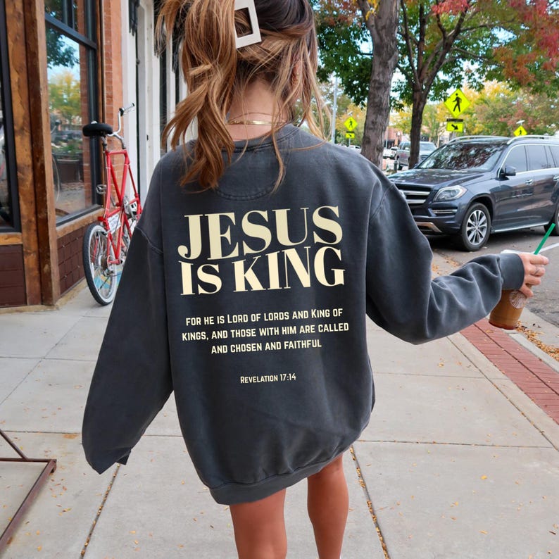 May include: Dark gray sweatshirt with the text "JESUS IS KING" in a bold, cream-colored font. Below the main text is a quote from Revelation 17:14. The sweatshirt is a casual style, suitable for everyday wear.
