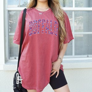 May include: A red t-shirt with the word "BUFFALO" in large, blue and white patterned letters across the chest. The shirt has short sleeves and a slightly faded appearance. The model is wearing black shorts and a black bag.