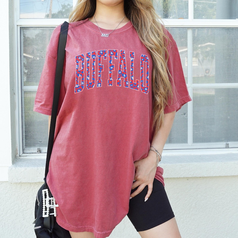 May include: A red t-shirt with the word "BUFFALO" in large, blue and white patterned letters across the chest. The shirt has short sleeves and a slightly faded appearance. The model is wearing black shorts and a black bag.