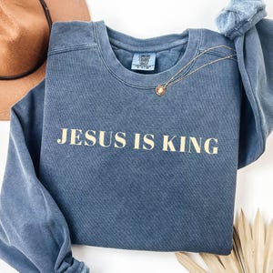 May include: A blue crewneck sweatshirt with the text "JESUS IS KING" in a light tan color. A gold necklace with a pendant is draped across the sweatshirt. A brown hat and a blue scrunchie are in the background.