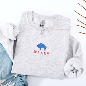 May include: A light gray sweatshirt with a blue buffalo graphic and the words "let's go" in red. A silver necklace with a small pendant is draped across the sweatshirt. Rolled up sleeves and a pair of blue jeans are visible.