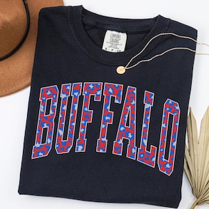 May include: Navy blue Comfort Colors t-shirt with the word "BUFFALO" in red, blue, and white leopard print lettering. A gold necklace with a small pendant is draped across the shirt. A brown hat is visible in the upper left corner.